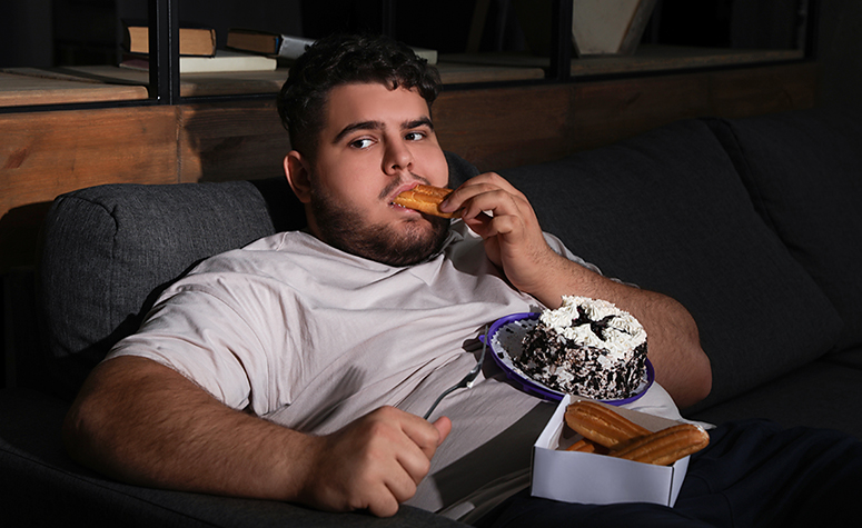 Food addict sat on sofa eating chocolate cakes