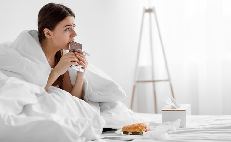 Woman sat in bed suffering from food addiction with chocolate and a burger