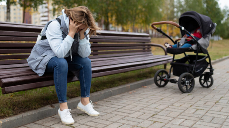Young woman sat on park bench suffering with postnatal depression with baby in pushchair
